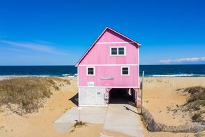 A pink beach house with a single door and a small porch, located on a sandy beach with the ocean in the background.