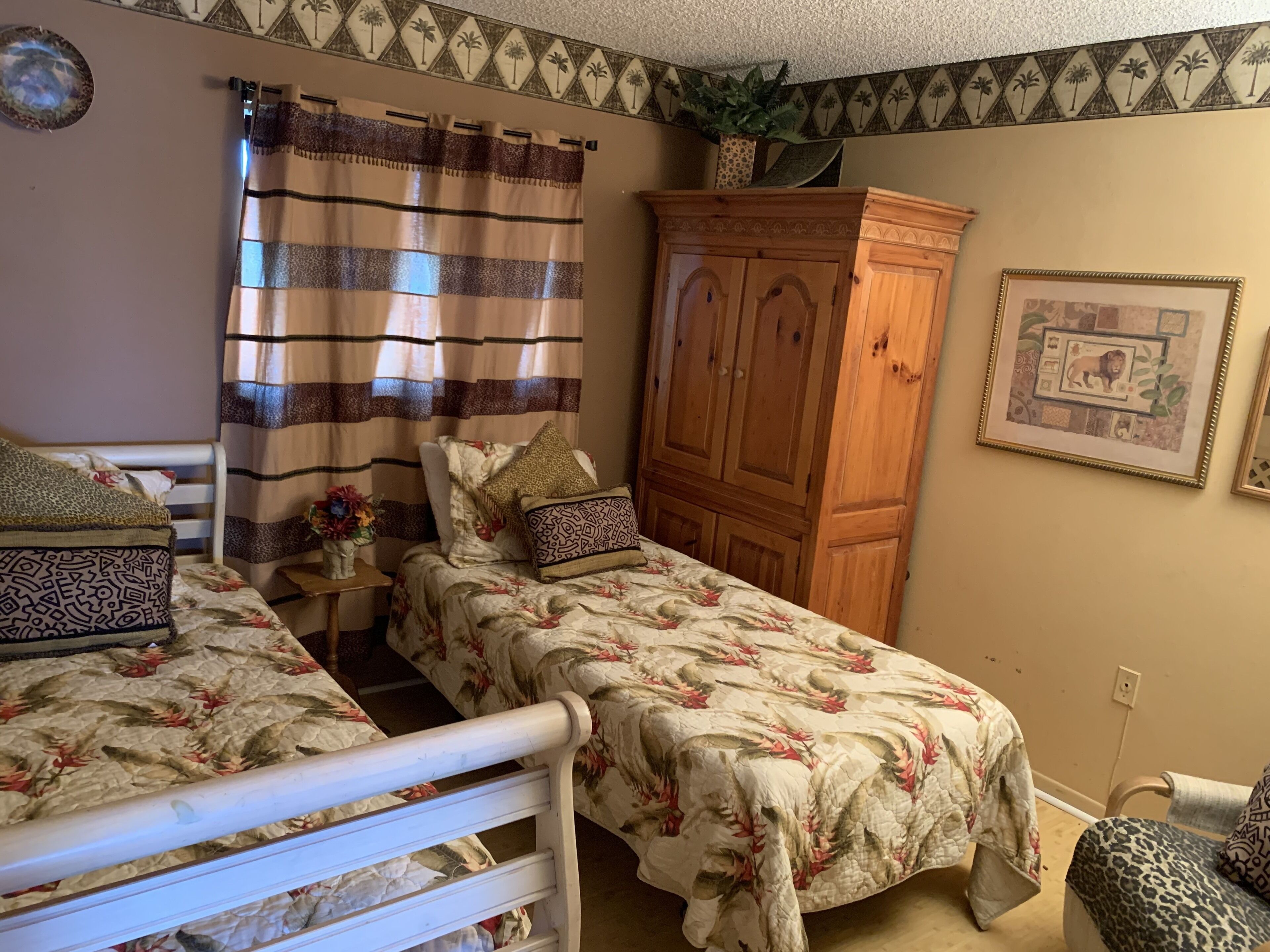 A bedroom with two single beds, a wooden wardrobe, a small table with a vase, and a framed picture on the wall.