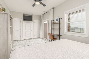 Une chambre à coucher avec un lit, un ventilateur de plafond, une fenêtre avec des stores et une vue sur les maisons à l’extérieur.