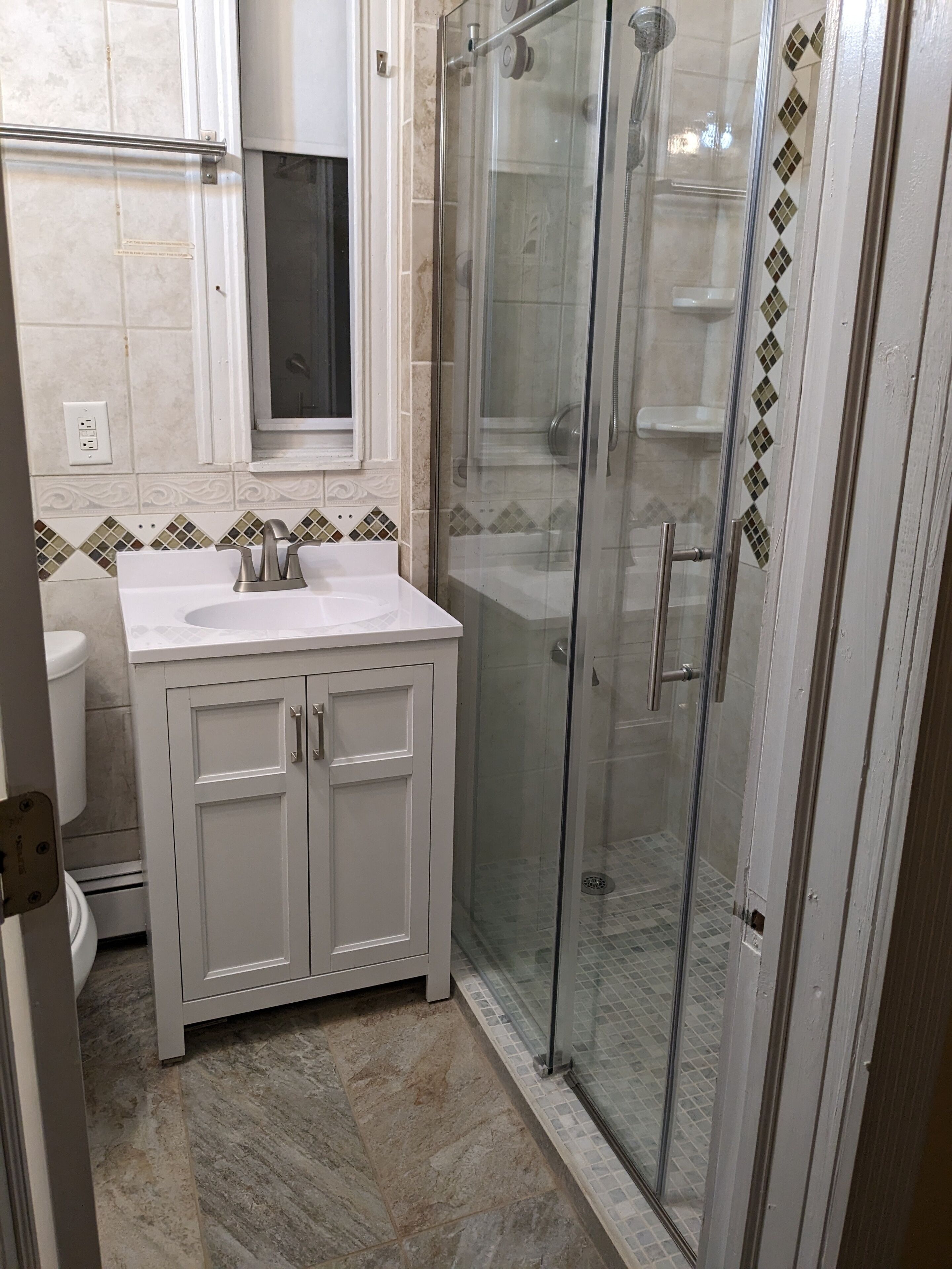 Een badkamer met een douche met glazen wand, een witte wastafel met een spiegel erboven en decoratieve tegels op de muren.