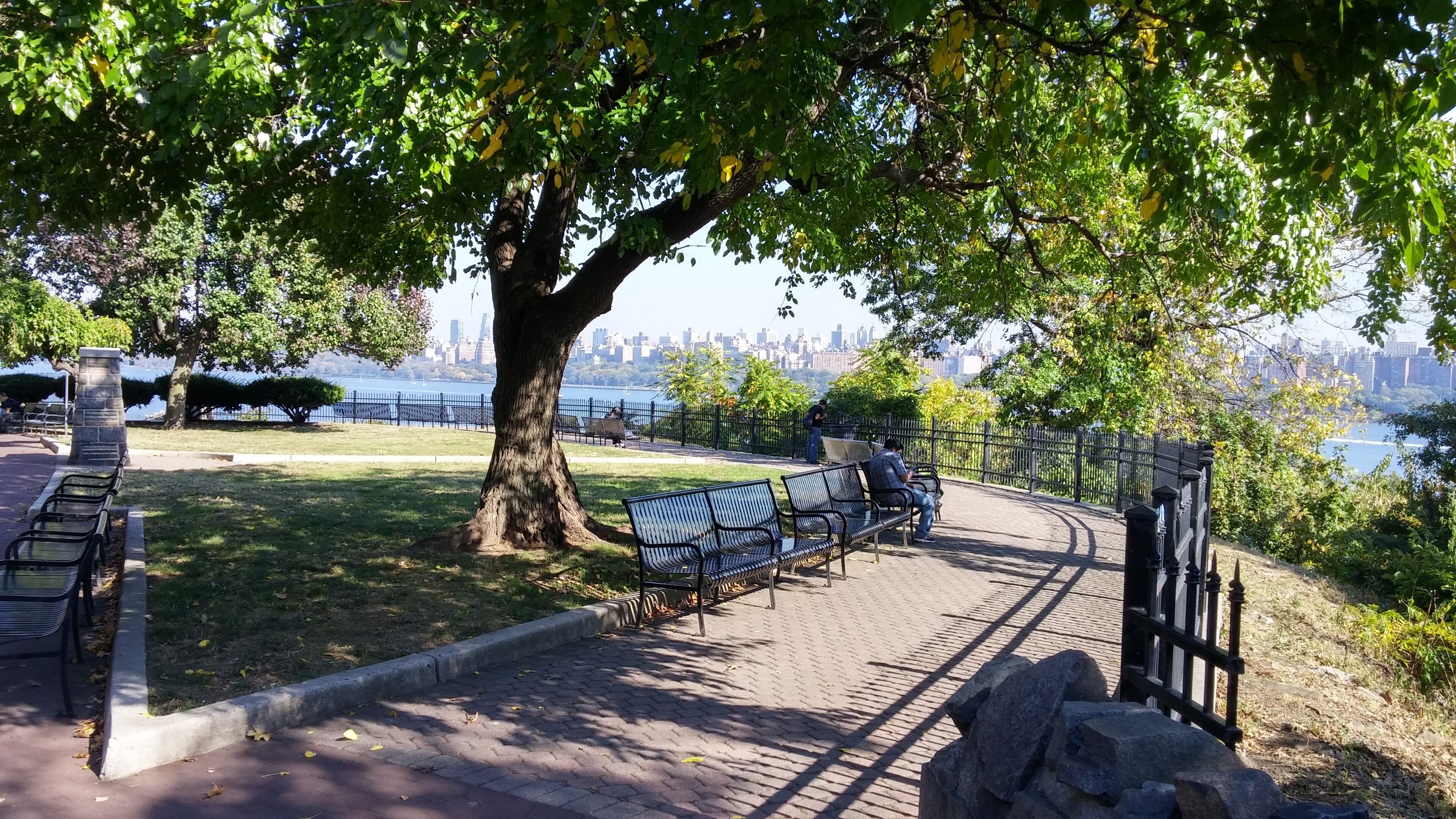 Ein Park mit Bänken, einer Steinsäule und Blick auf eine Stadtlandschaft über dem Wasser.
