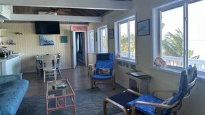 A cozy living room with a wooden ceiling, a television mounted on the wall, and a view of the outdoors through large windows. - Beachfront large 2/2 on Spanish Wells' northside beach. Sun deck by the water. (Spanish Wells)