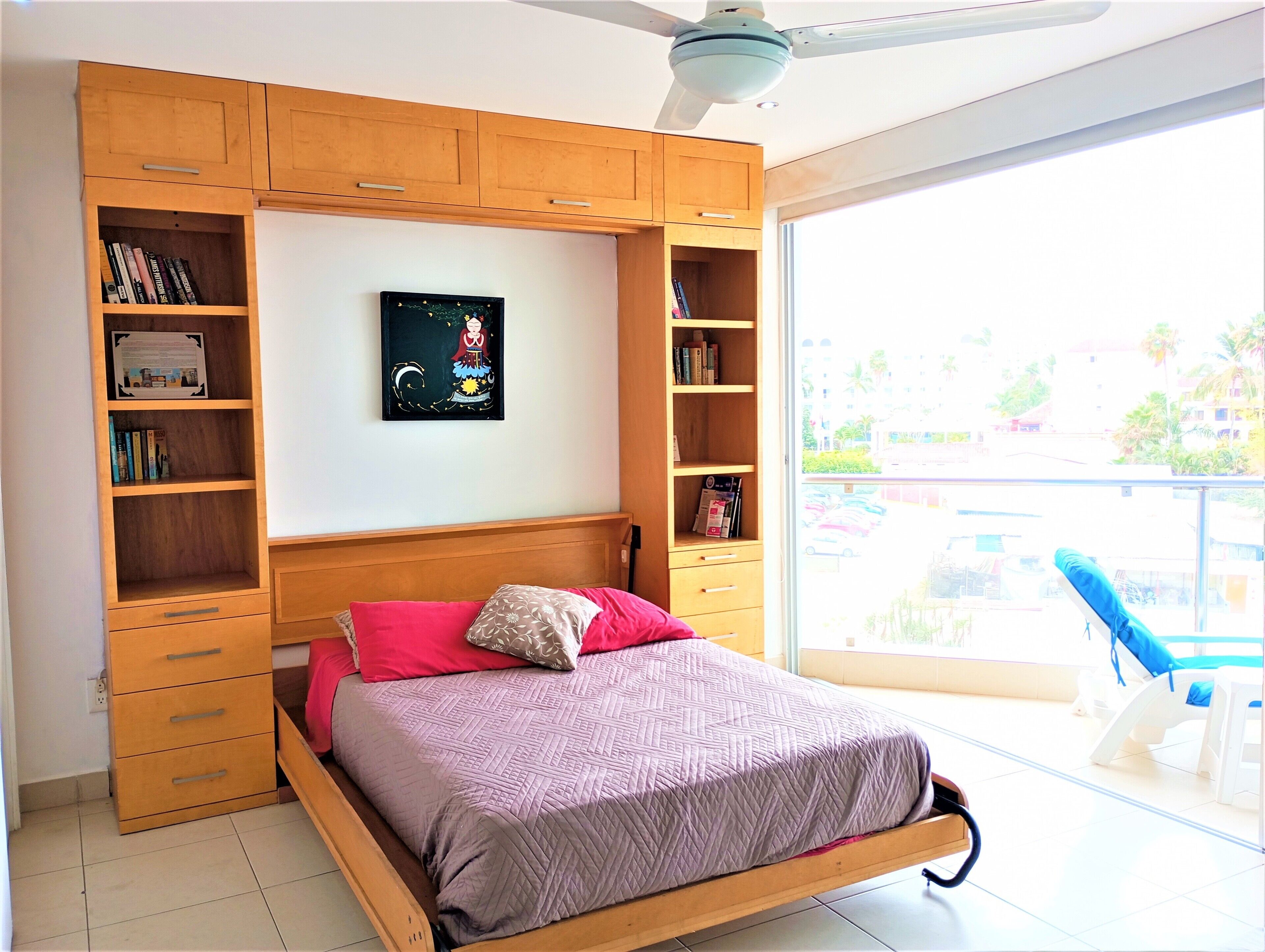 A bedroom with a bed, wooden shelving, a ceiling fan, and a view of the outside.