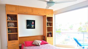 A bedroom with a bed, wooden shelving, a ceiling fan, and a view of the outside.