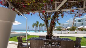 Een overdekt terras met rieten meubels, een tafel met flessen, en zicht op een zwembad en strand in de verte.
