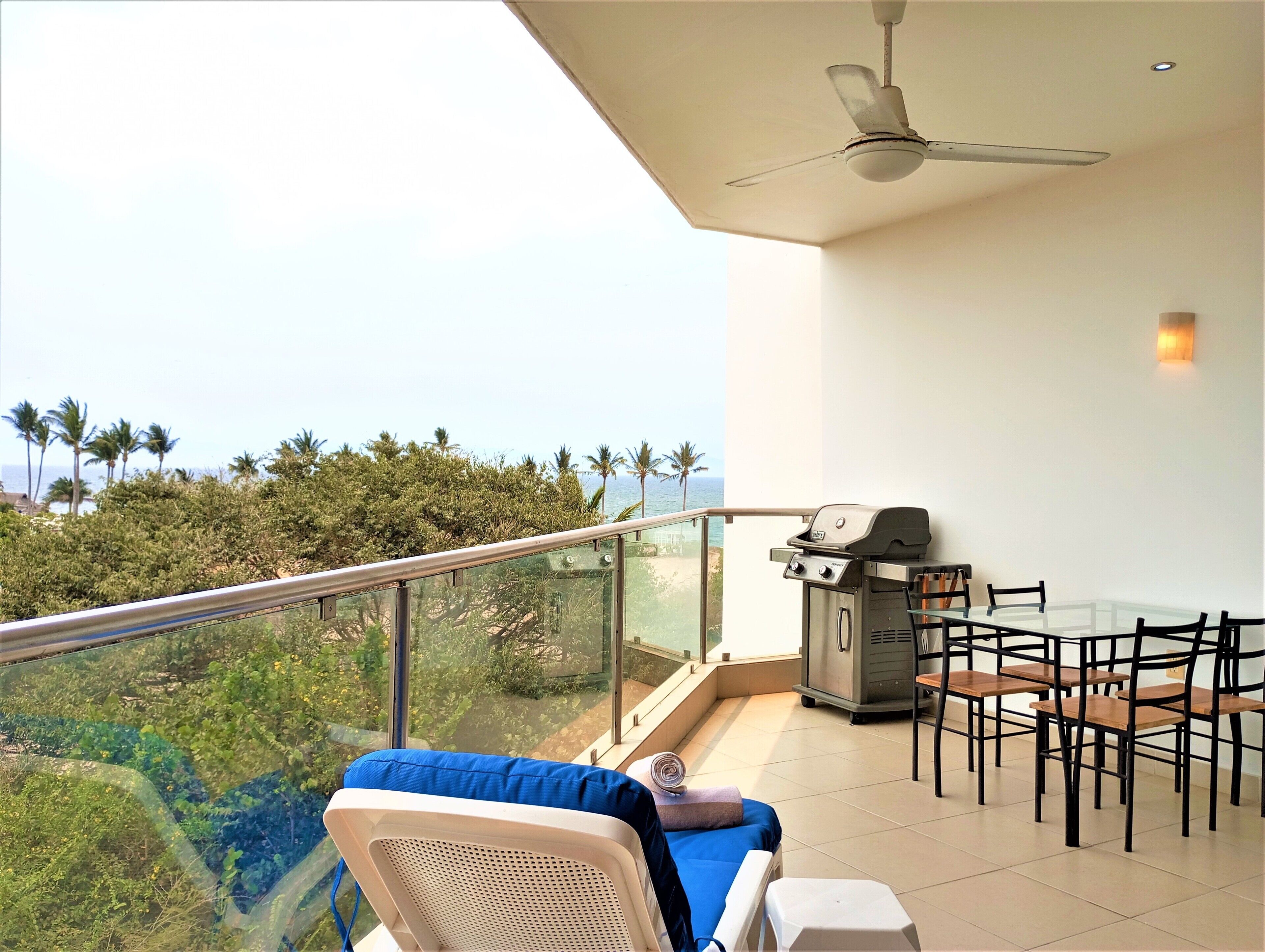 A balcony with a glass railing, a ceiling fan, a grill, and a table with chairs.