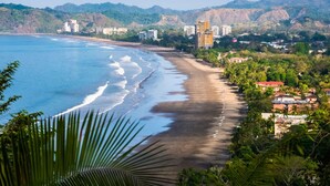 A coastal view with a beach, buildings, and hills in the background. - Hotel Tropical Garden (Jacó)