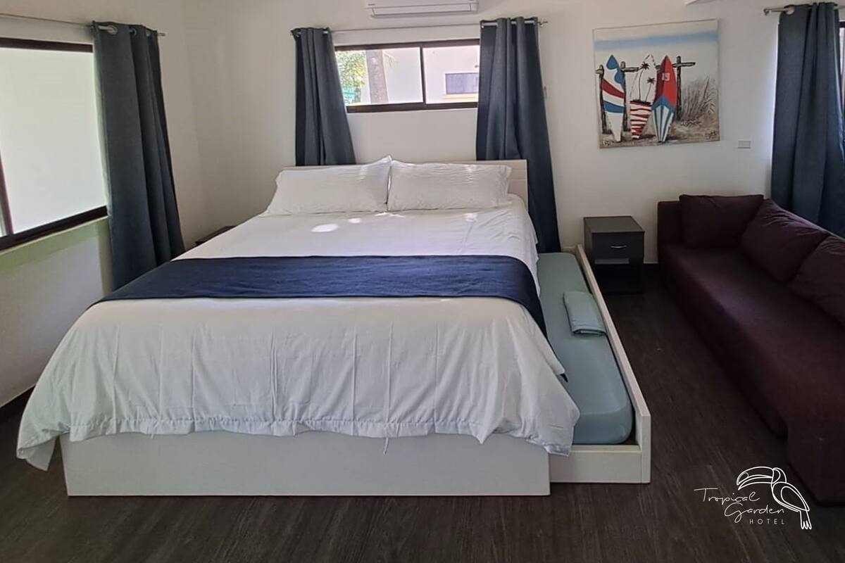A bedroom with a bed, a sofa, an air conditioning unit, a window, and a painting on the wall.