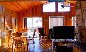 Cozy cabin-style room with a large open-plan layout, dining area, and seating area, featuring a fireplace and large windows.