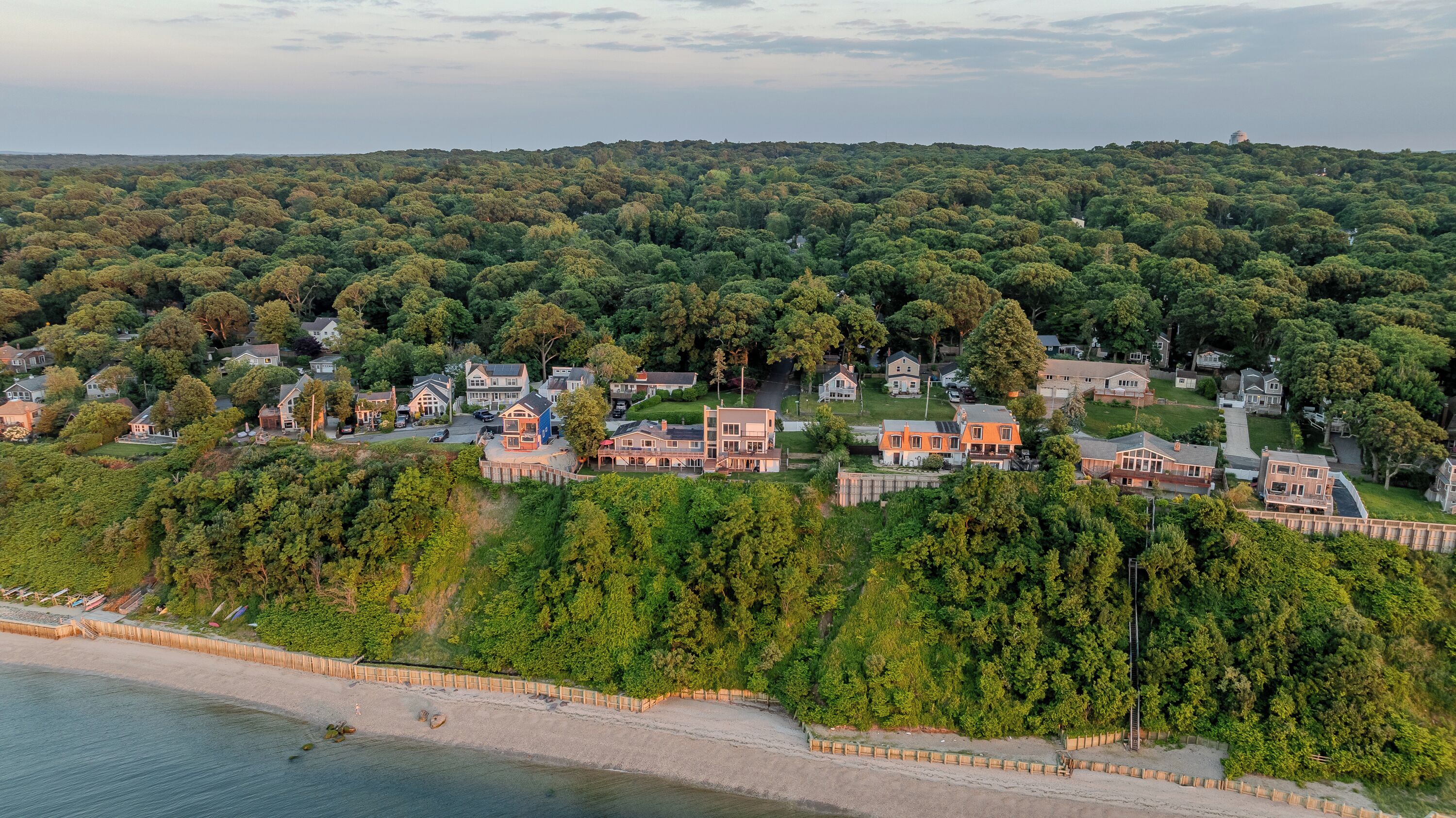 Vista aérea de una zona residencial con casas rodeadas de un bosque denso, una playa y un faro a lo lejos.