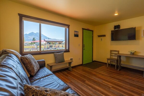 A room with a wooden floor, a sofa, a bench, a green door, and a TV mounted on the wall.