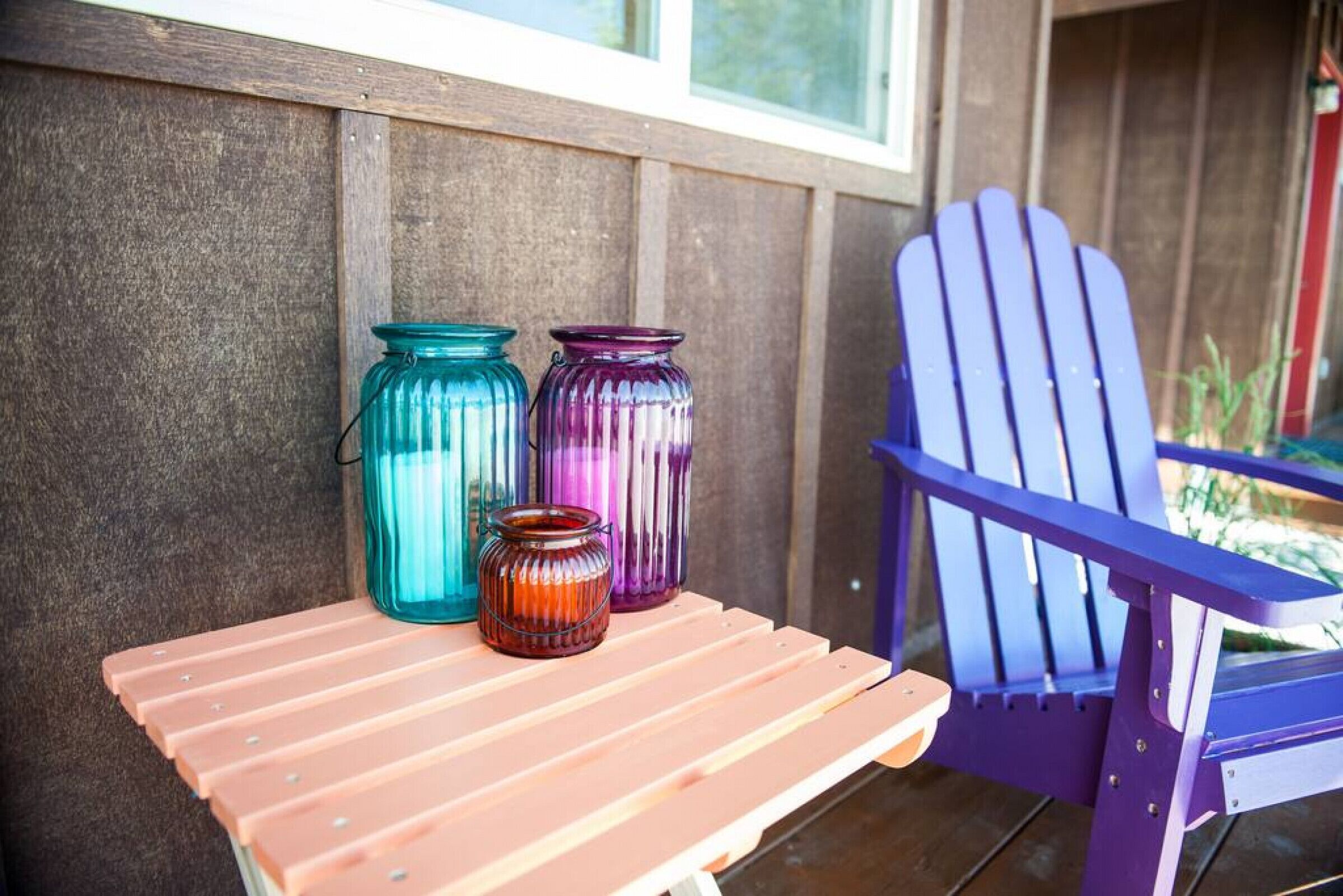 A wooden table with three glass jars and a purple Adirondack chair on a porch.