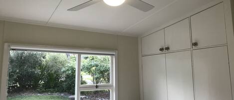 A room with a ceiling fan, a window with a view of a garden, and a wardrobe with multiple doors.