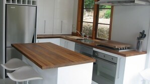 Een kleine keuken met witte kastjes, een roestvrijstalen koelkast, een houten aanrechtblad en een raam met uitzicht op het landschap.
