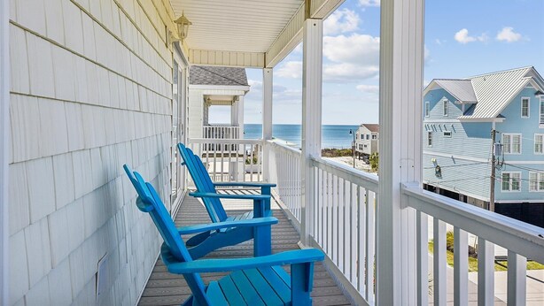 A covered porch with blue Adirondack chairs, a white railing, and a view of the ocean and beach houses.