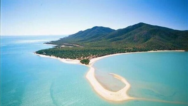 Vista aérea de una zona costera con una playa arenosa curvada, aguas turquesas cristalinas y una montaña boscosa al fondo.