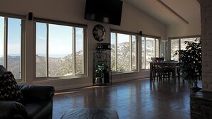 A spacious living room with large windows, a ceiling fan, a flat-screen TV, and a dining area. - Quiet Hilltop Home With Stunning Hill Top And Valley Views (Valley Center)