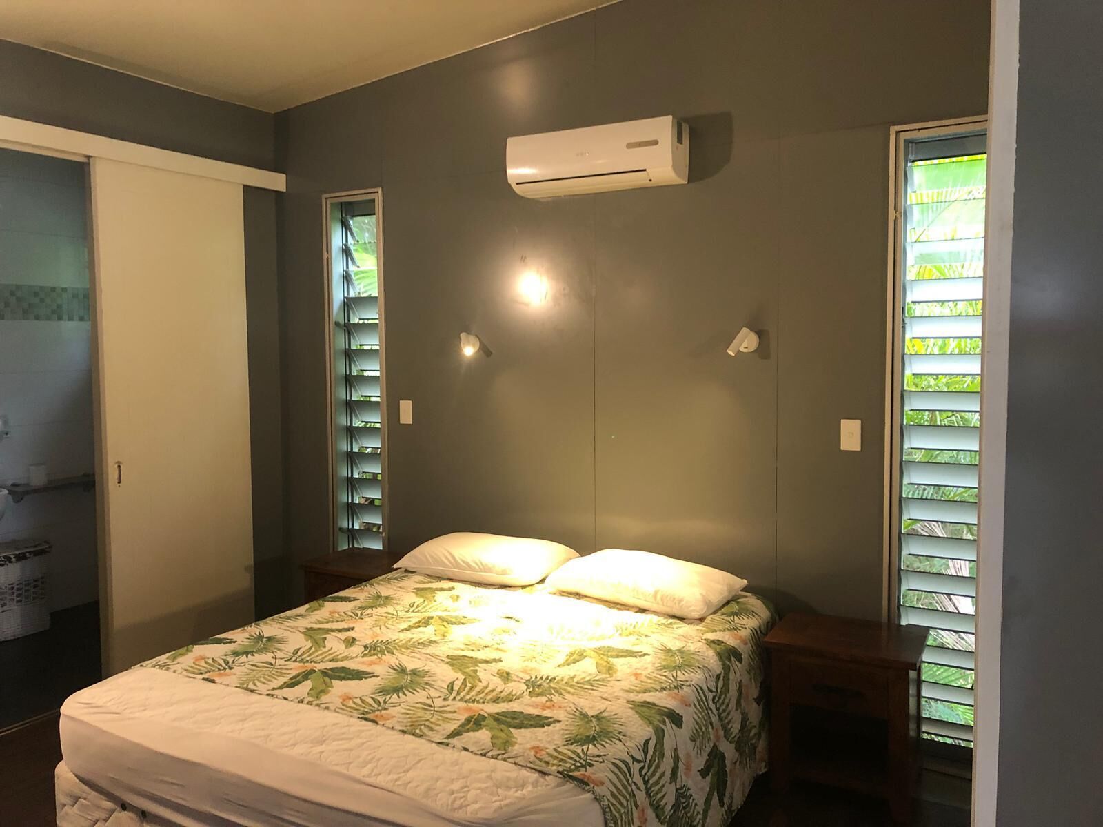 Quarto com cama, ar condicionado, janela com persianas e porta que dá para um banheiro.