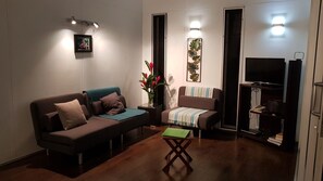 A modern living room with a sofa, armchair, and a small table. There is a TV on a stand and a bookshelf with books. - Premier and Stunning Beach Front Location UNIT 4 (Nikao)