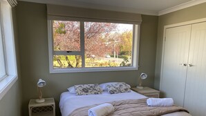 A neatly made bed with a beige quilt, two bedside lamps, and a window with a view of trees. -  Located near Eely Point  (Wanaka)