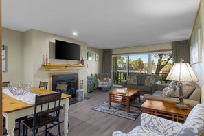A living room with a fireplace, a television, a dining area with a table and chairs, and a sofa with a view of the outdoors.