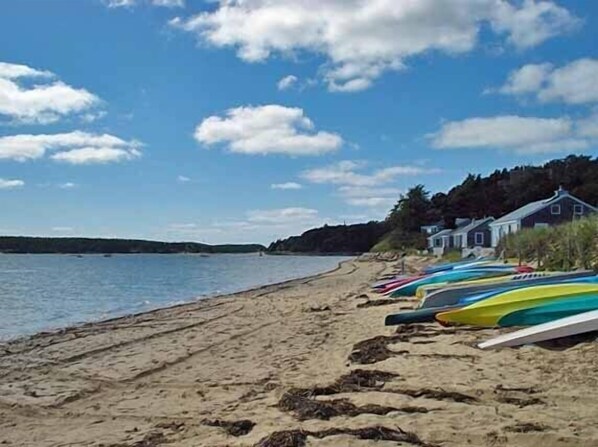 A beach with several colorful kayaks lined up on the sand, a calm sea, and a few houses in the background.