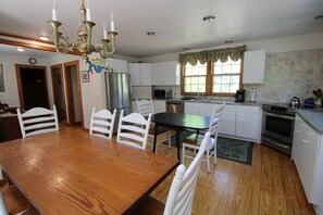 A kitchen with white cabinets, a stainless steel refrigerator, a microwave, and a stove. There is a dining table with chairs and a chandelier. - Private & Spacious 5 Bedroom Home (1607) (Wellfleet)