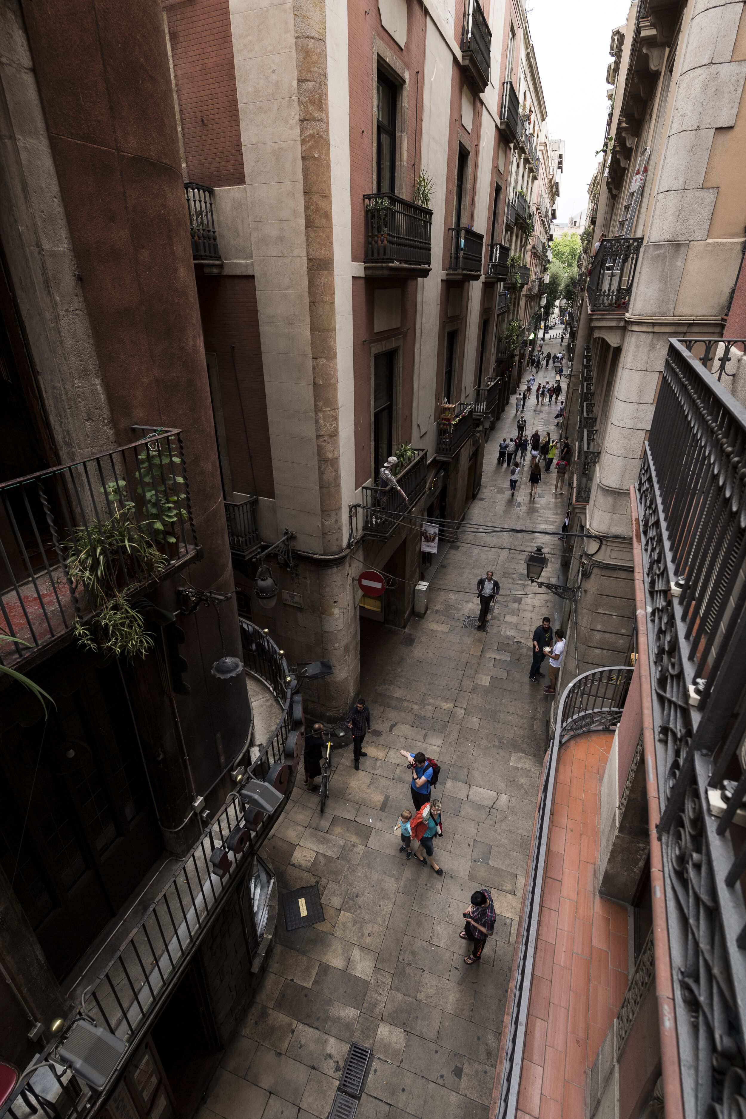 A narrow urban street with buildings on both sides, featuring balconies, a few pedestrians, and a motorbike.