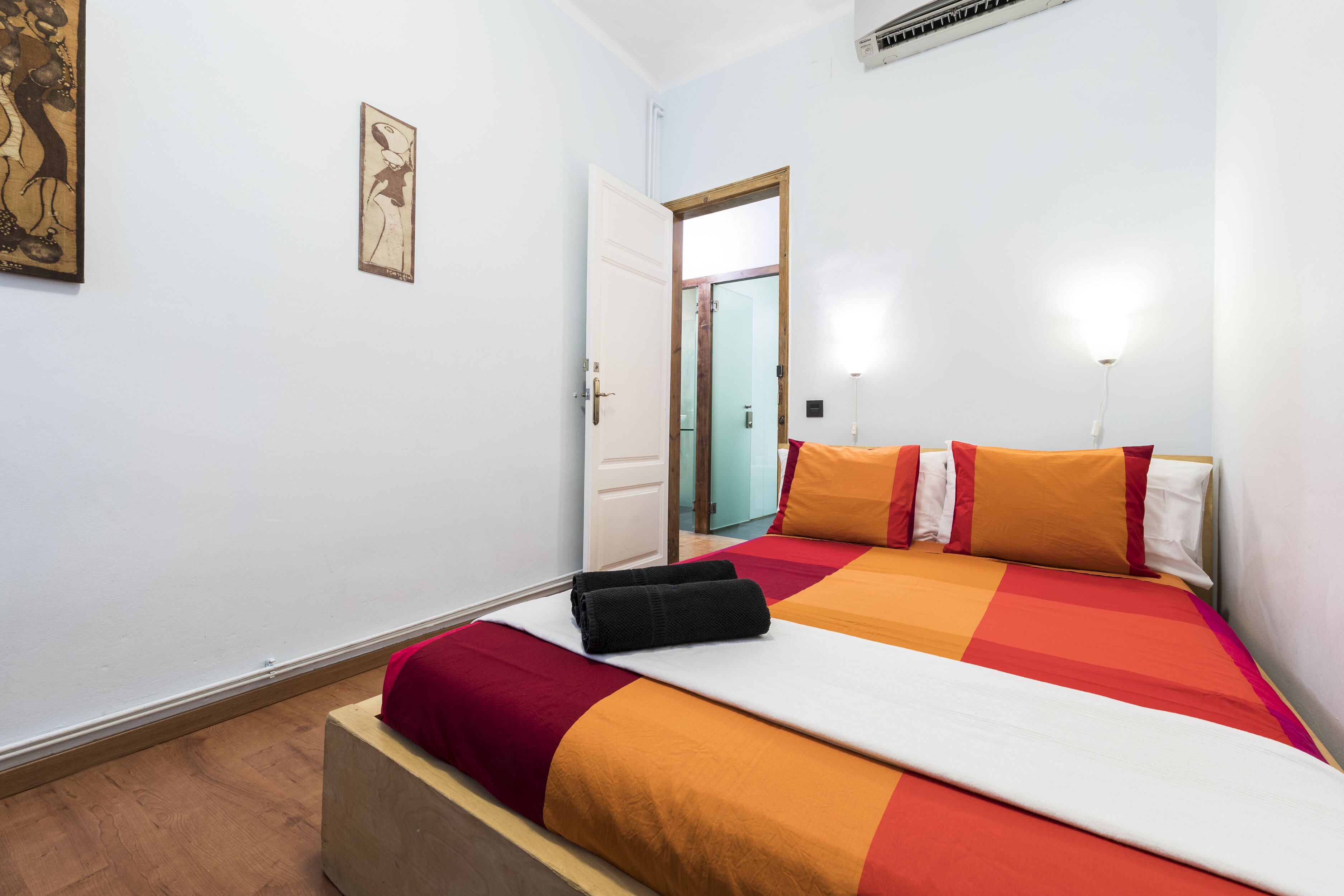 A neatly made bed with a red and orange bedspread, a wooden bedside table, and a door leading to another room.