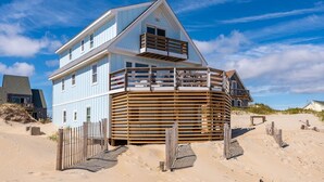 A coastal house with a prominent balcony, a sandy beach in the background, and a clear blue sky. - 600 Nags Head Ocean-Front Completely Renovated (Nags Head)