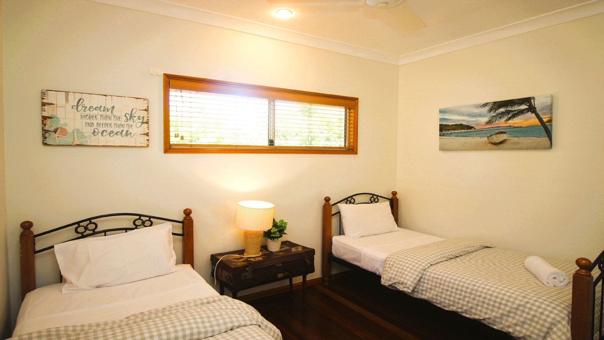 A room with two single beds, a nightstand with a lamp, a window with blinds, and wall art.