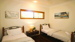 A room with two single beds, a nightstand with a lamp, a window with blinds, and wall art.