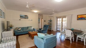 Ein Wohnzimmer mit einer Couch, Sesseln, einem Couchtisch, einem Esstisch mit Stühlen, Deckenventilatoren und einem Bücherregal.
