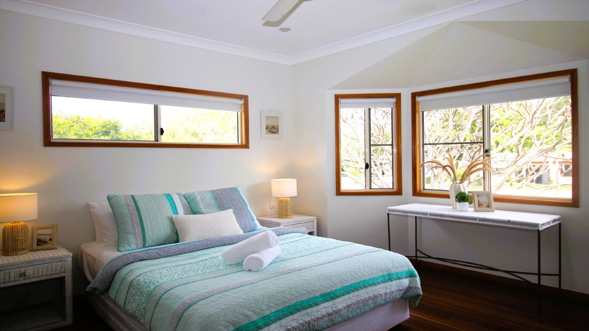 A bedroom with a bed, nightstands, a table, and windows with views of greenery.
