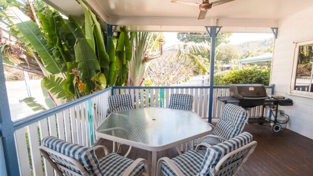 Eine überdachte Terrasse mit einem Glastisch, umgeben von gestreiften Stühlen, einem Grill und üppigen tropischen Pflanzen.