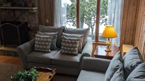 Una sala de estar acogedora con un sofá gris, una mesa de centro de madera y una lámpara sobre una mesita auxiliar. Se ve una pared de piedra y una ventana con cortinas.