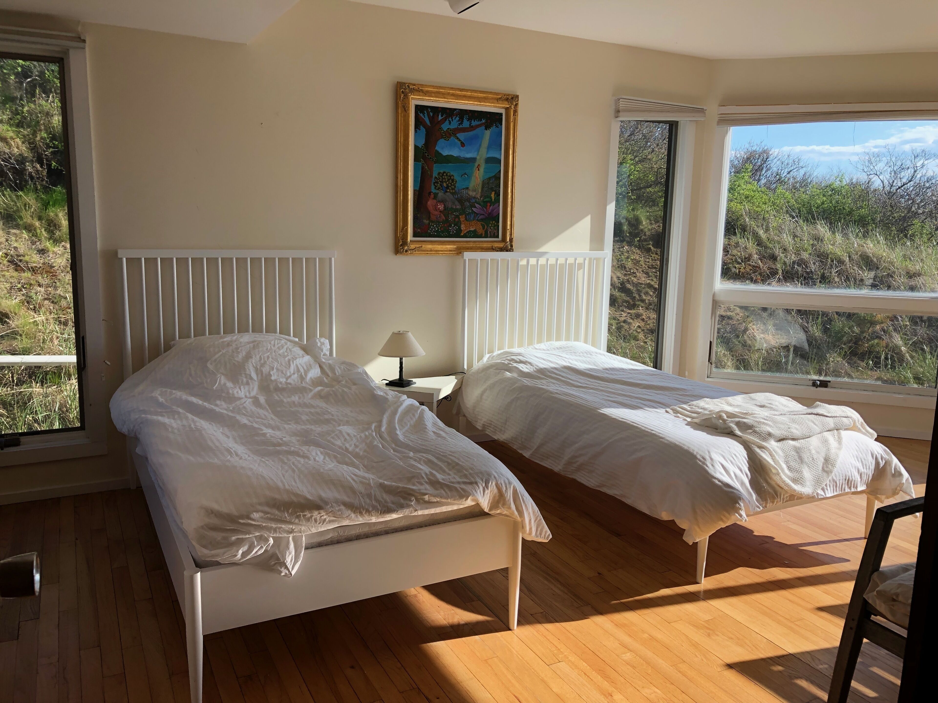 A bedroom with two beds, a painting on the wall, and large windows with a view of the outdoors.