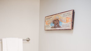 Et badeværelse med trægulv, hvide vægge, et hvidt håndklæde hængende på en metalhåndklædestang og en vintageplakat på væggen.