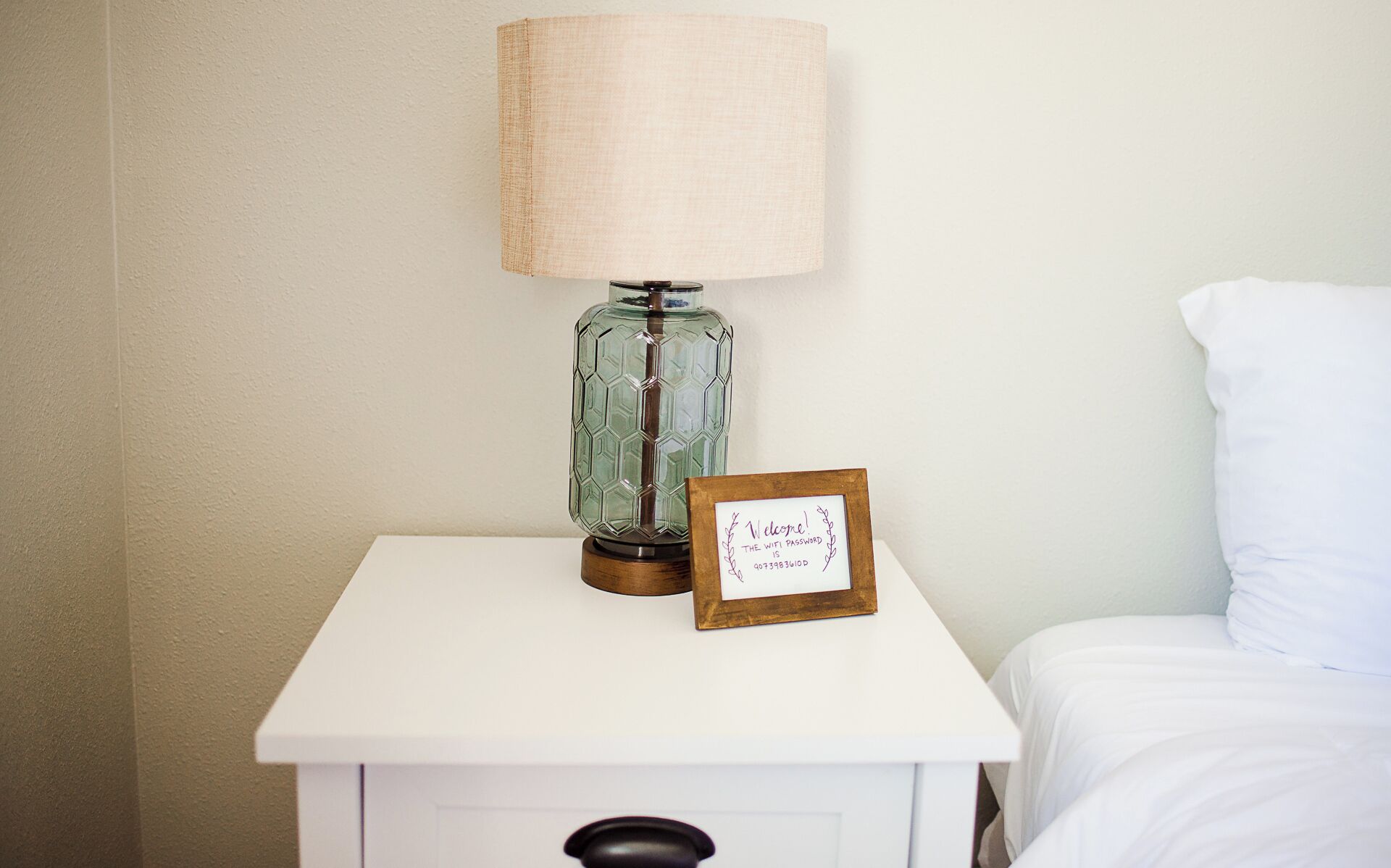 A bedside table with a lamp and a framed welcome sign.