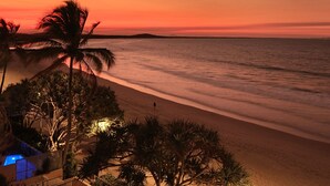 A beach at sunset with palm trees and a pool area.