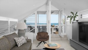 A modern living room with a sofa, a chair, a small table, and a TV. There is a staircase leading to an outdoor area with a view of the sea.