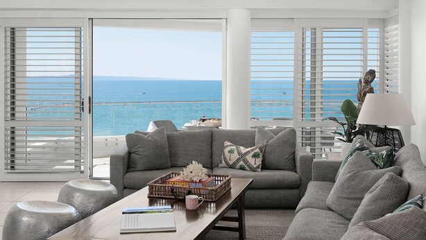 A living room with a large window overlooking the sea, a sofa set, a coffee table with books and a decorative basket, and a lamp.