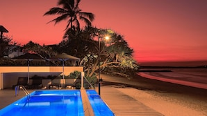 A swimming pool with a wooden deck, a palm tree, and a beach in the background.