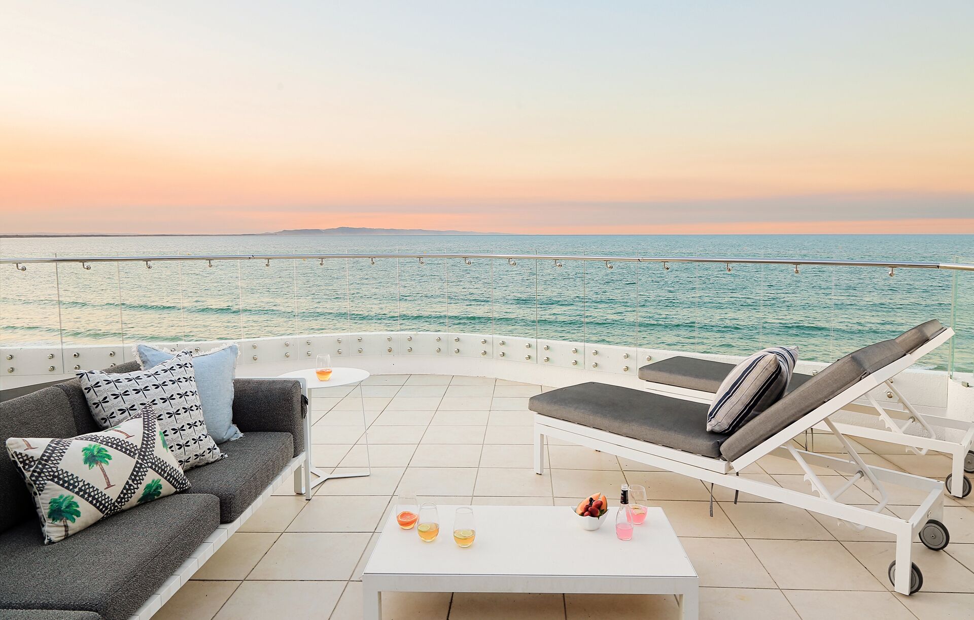 A sunlit terrace with a view of the sea, a sofa, lounge chairs, and a table with drinks and fruit.