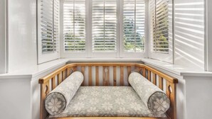 Een erker met witte luiken, een houten bank met gekleurde kussens, en uitzicht op groen.