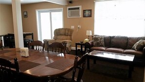 A living room with a dining table, chairs, a sofa, and a coffee table. There is a window with blinds, an air conditioning unit on the wall, and a door. - Visiting Cooperstown NY or Oneonta NY areas stay at Abel's Acre's Hideaway House (Mt Vision)