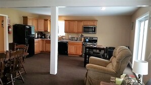 A spacious living room with a beige armchair and a dining area with wooden chairs. A kitchen with wooden cabinets and modern appliances is visible in the background. - Visiting Cooperstown NY or Oneonta NY areas stay at Abel's Acre's Hideaway House (Mt Vision)