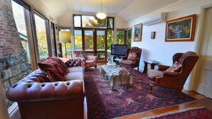 A cozy living room with a leather sofa, armchairs, and a television. Large windows offer a view of trees outside. - ONLY PROPERTY IN THE VILLAGE WITH A POOL AND PRIVATE RIVER FRONTAGE (Kangaroo Valley)