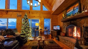 A cozy cabin interior with a Christmas tree, leather sofa, stone fireplace, and a television.