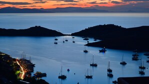 Una marina costiera al crepuscolo con barche ancorate in acque calme, circondata da colline.